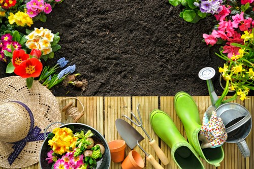 A vibrant, well-organized back garden scene showing a wooden decking area bordered by a layer of dark, freshly turned soil. Along the top edge, there are colorful flower beds filled with bright pink, purple, yellow, and red flowering plants and shrubs. On the decking, various gardening tools and accessories are neatly arranged, including a pair of bright green rain boots, a metal watering can with a floral design, small terracotta pots, a trowel, and a hand rake. A wide-brimmed straw hat with a purple ribbon is also placed on the decking, suggesting outdoor gardening activity. To the left, a black plastic container holds a small, lush arrangement of pink, yellow, and purple flowers, adding natural colour contrasts to the setting. The environment appears to be part of a well-maintained residential garden, suitable for outdoor gardening and landscaping projects, with an emphasis on vibrant plant colour and organized outdoor tools typical of professional gardening services in Elephant and Castle.